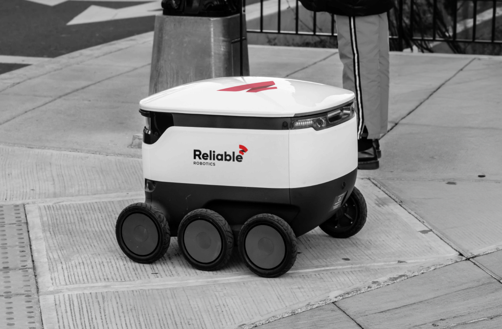 Black and white photo of a Reliable Robotics autonomous delivery robot on a city sidewalk, featuring the new red logo.
